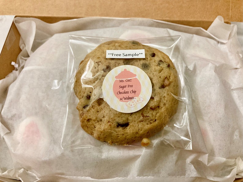 May include: A clear plastic bag containing a single sugar-free chocolate chip cookie with walnuts. The cookie has a pink and yellow label that reads "Ms. Geez Sugar Free Chocolate Chip w/Walnuts". The bag has a white label that reads "**Free Sample**".