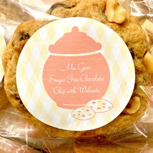 May include: A close-up of a sugar-free chocolate chip cookie with walnuts. The cookie is in a clear plastic bag with a yellow and white label that says "Ms. Gooz Sugar Free Chocolate Chip with Walnuts".