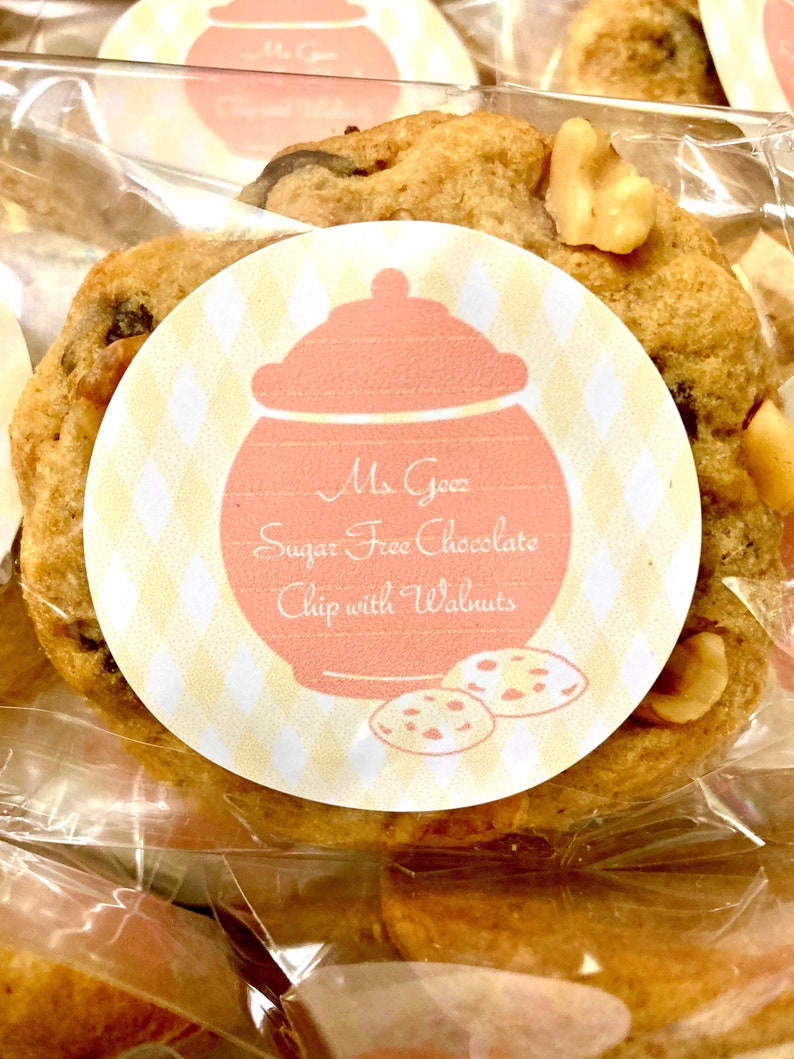 May include: A close-up of a sugar-free chocolate chip cookie with walnuts. The cookie is in a clear plastic bag with a yellow and white label that says "Ms. Gooz Sugar Free Chocolate Chip with Walnuts".