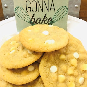 May include: A close-up of a stack of freshly baked cookies with white chocolate chips. The cookies are golden brown and have a soft, chewy texture. The sign behind the cookies reads "Bakers gonna bake".