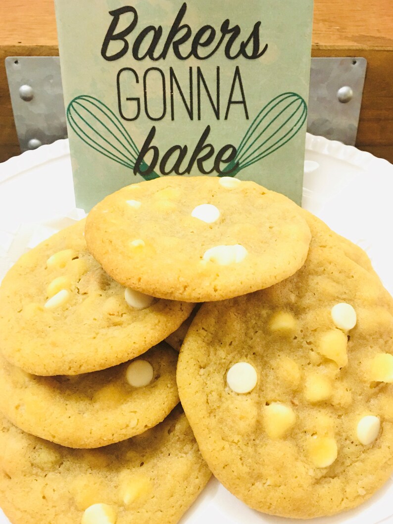 May include: A close-up of a stack of freshly baked cookies with white chocolate chips. The cookies are golden brown and have a soft, chewy texture. The sign behind the cookies reads "Bakers gonna bake".