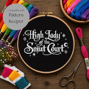 May include: A round wooden embroidery hoop containing black fabric with white cross-stitched text that reads High Lady of the Smut Court, surrounded by colorful embroidery floss, a pair of metal scissors, and a small bouquet of flowers.