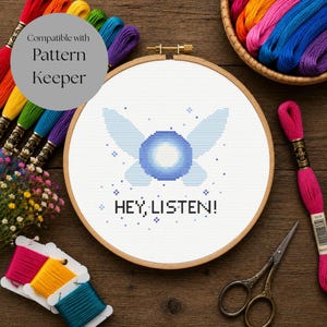 May include: A circular wooden embroidery hoop containing a cross-stitch design of a glowing blue fairy with wings and the text HEY, LISTEN! surrounded by small blue stars, accompanied by colorful embroidery floss, scissors, and small flowers.