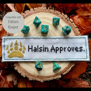 May include: A white cross-stitch banner with a gold paw print and the text "Halsin Approves."  The banner is surrounded by green and gold dice on a wooden surface.