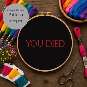 May include: A round wooden embroidery hoop containing black fabric with the red stitched text YOU DIED. The hoop is surrounded by colorful embroidery floss skeins, a small pair of metal scissors, and a small bouquet of dried flowers.