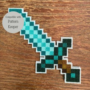 May include: A pixelated sword in teal, green, and brown, with a white outline. The text "Compatible with Pattern Keeper" is in a grey circle in the upper left corner. The sword is on a wooden surface.