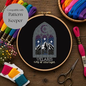 以下が含まれることがあります： 木製の刺繍枠に入ったクロスステッチ作品。黒い背景に灰色のアーチ型窓枠、三日月、星、雪を頂いた山々が描かれています。テキストは「VELARIS city of starlight」と記されています。刺繍糸とハサミが含まれています。
