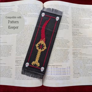 May include: Black bookmark with a gold key and red flame design, accented with white skulls. The bookmark is made of fabric and has a fringed edge. The text "Compatible with Pattern Keeper" is in the upper left corner.