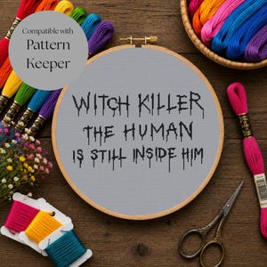 May include: A circular wooden embroidery hoop containing gray Aida fabric with black cross-stitched text that reads Witch Killer The Human Is Still Inside Him. The hoop is surrounded by colorful embroidery floss, a pair of metal scissors, and flowers.