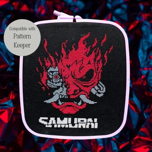 May include: Cross-stitch embroidery featuring a red oni mask design with the word "SAMURAI" in white. The design is set against a black background within a square, light purple frame. The text "Compatible with Pattern Keeper" is in a circle in the upper left.