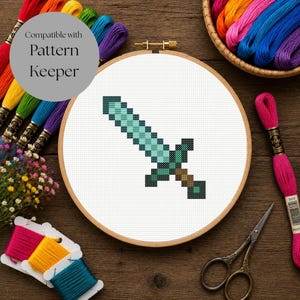 May include: A circular wooden embroidery hoop containing a cross-stitch pattern of a pixelated sword in shades of light blue, dark blue, and brown. The design is set against a white fabric background, surrounded by colourful embroidery floss and scissors.