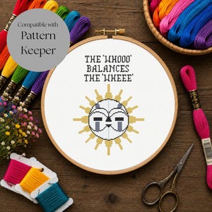 May include: A cross stitch project in a wooden embroidery hoop featuring a stylized sun graphic with a face and the text THE 'WHOOO' BALANCES THE 'WHEEE'. The design is on white fabric, surrounded by colorful embroidery floss and scissors.