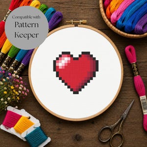 Op de afbeelding: Een houten borduurring met witte stof waarop een pixelachtig rood hart in kruissteek is geborduurd. De tekst Compatible with Pattern Keeper is zichtbaar. Rondom de ring liggen kleurrijke strengen borduurgaren en een schaar.
