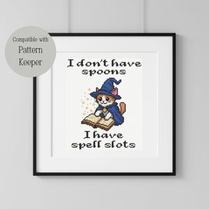 May include: Framed cross-stitch art depicting a cat wizard in a blue hat and cape, reading a spell book. The text reads "I don't have spoons, I have spell slots." The artwork is in a black frame with a white mount.
