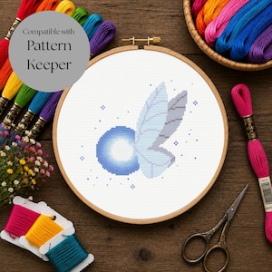 Peut inclure: Un tambour à broder circulaire en bois contenant un motif au point de croix représentant un orbe bleu lumineux avec des ailes bleu clair et des étoiles. Le texte Compatible with Pattern Keeper est visible. Fils et ciseaux sont présents.