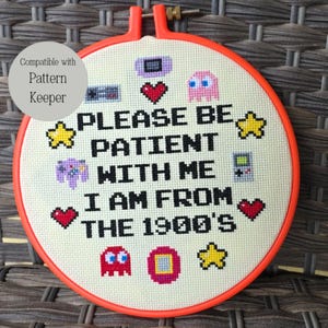 May include: A cross-stitch hoop with the text "PLEASE BE PATIENT WITH ME I AM FROM THE 1900'S" and pixelated game graphics. The hoop is orange, and the fabric is off-white. The text is in black.