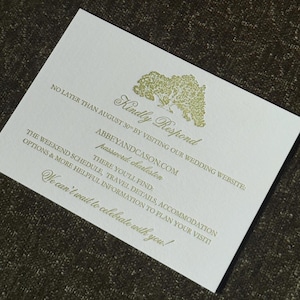 May include: A white invitation card with gold text and a tree illustration. The text includes "Kindly Respond," a website address, and other wedding details. The card is on a dark, textured surface.