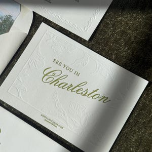 以下が含まれることがあります： エンボス加工された花柄のデザインと、緑色の「See You in Charleston」の文字が入った白い招待状。カードには「2025年10月」とウェブサイト「ABBEYANDCASON.COM」も記載されています。