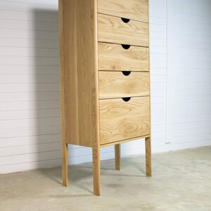 May include: A tall, light wood chest of drawers with six drawers and black drawer pulls. The chest of drawers has four legs and is standing on a concrete floor.