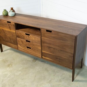 Puede incluir: Un aparador de madera de nogal con cuatro cajones y dos puertas. El aparador tiene un dise&ntilde;o moderno con l&iacute;neas limpias y un acabado de madera natural. Est&aacute; sobre un suelo de hormig&oacute;n.