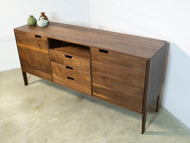 Puede incluir: Un aparador de madera de nogal con cuatro cajones y dos puertas. El aparador tiene un dise&ntilde;o moderno con l&iacute;neas limpias y un acabado de madera natural. Est&aacute; sobre un suelo de hormig&oacute;n.