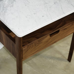 May include: A wooden nightstand with a white marble top and a drawer. The nightstand has a simple design with straight lines and a small shelf.