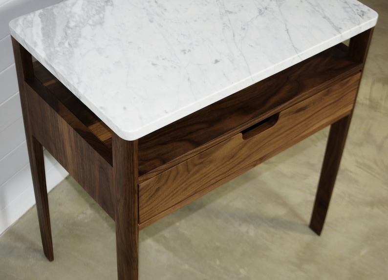 May include: A wooden nightstand with a white marble top and a drawer. The nightstand has a simple design with straight lines and a small shelf.