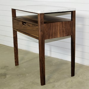May include: A walnut wood side table with a white marble top and a drawer. The table has a simple, modern design with clean lines and a minimalist aesthetic.