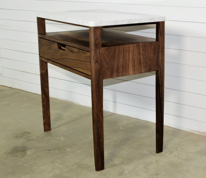 May include: A walnut wood side table with a white marble top and a drawer. The table has a simple, modern design with clean lines and a minimalist aesthetic.
