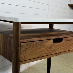 May include: A walnut wood end table with a white marble top. The table has a single drawer and an open shelf.