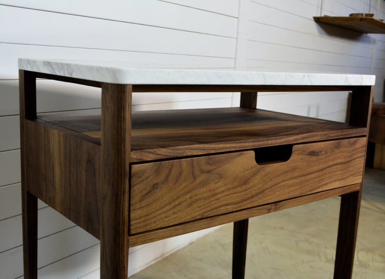 May include: A walnut wood end table with a white marble top. The table has a single drawer and an open shelf.