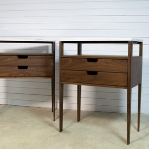 May include: Two matching walnut wood bedside tables with white marble tops and two drawers each. The bedside tables have a simple, modern design with thin legs.