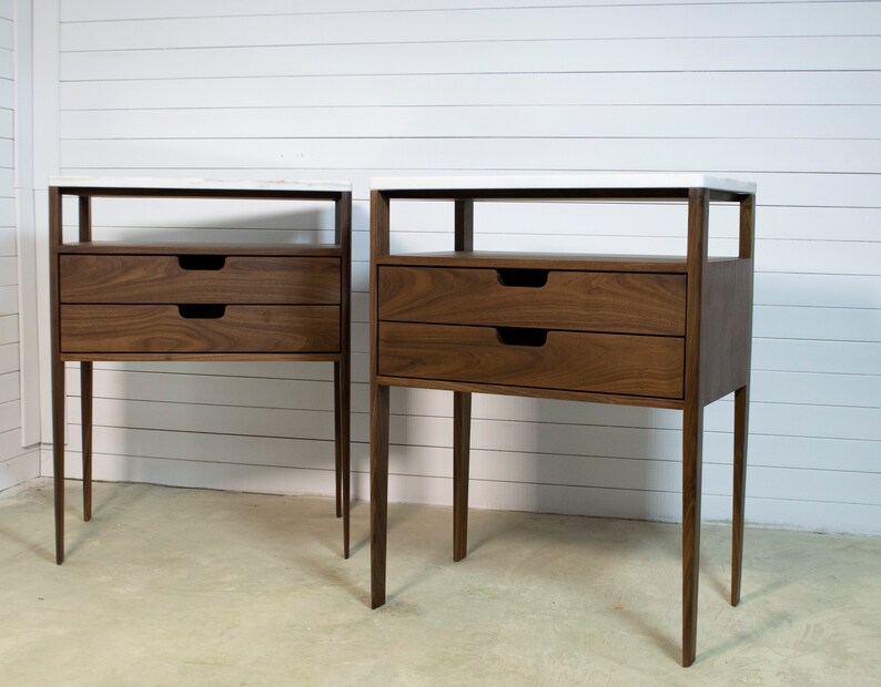 May include: Two matching walnut wood bedside tables with white marble tops and two drawers each. The bedside tables have a simple, modern design with thin legs.