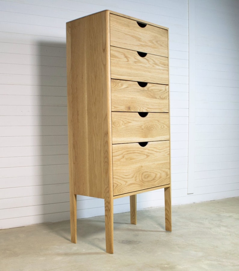 May include: A tall, light wood chest of drawers with six drawers and black drawer pulls. The chest of drawers has four legs and is standing on a concrete floor.