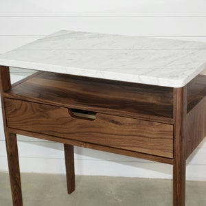 May include: A walnut wood end table with a white marble top. The table has a single drawer and a shelf below the top. The legs are tapered and the table has a modern design.