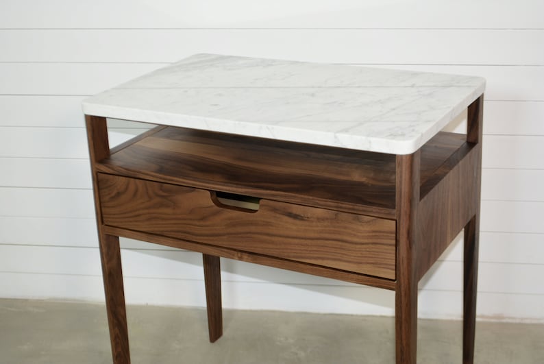 May include: A walnut wood end table with a white marble top. The table has a single drawer and a shelf below the top. The legs are tapered and the table has a modern design.