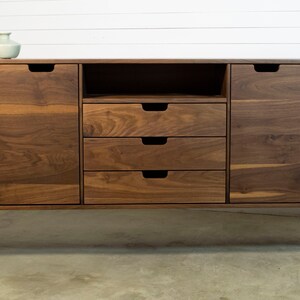 Puede incluir: Un aparador de madera de nogal con cuatro cajones y dos puertas. El aparador tiene un dise&ntilde;o moderno con patas de metal negro y un aspecto simple y limpio.