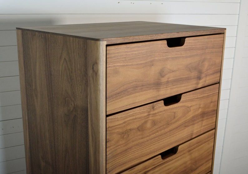 May include: A tall wooden dresser with five drawers. The dresser is made of a light brown wood with a natural finish. The drawers have simple, recessed handles.