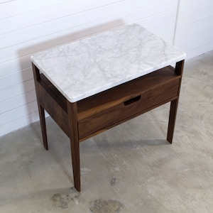 May include: A walnut wood nightstand with a white marble top and a single drawer. The nightstand has a simple, modern design and is perfect for a bedroom.