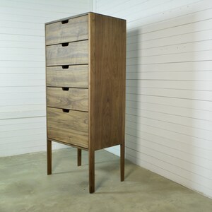 May include: A tall wooden dresser with five drawers and a dark brown finish. The dresser has four legs and is standing on a light brown floor.