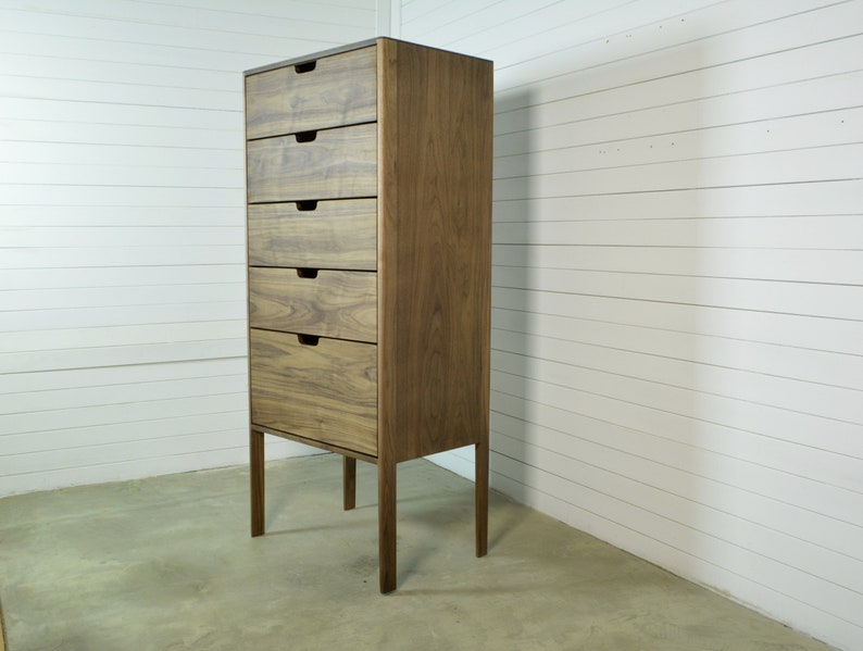 May include: A tall wooden dresser with five drawers and a dark brown finish. The dresser has four legs and is standing on a light brown floor.