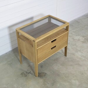 Puede incluir: Una mesa auxiliar de madera clara con dos cajones y una tapa de vidrio. La mesa tiene un dise&ntilde;o simple y moderno.