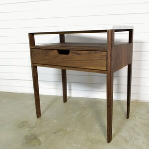 May include: A walnut wood end table with a single drawer and a shelf. The table has a modern design with clean lines and a simple, elegant look.