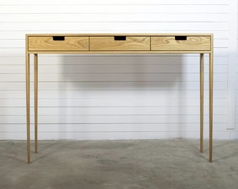 May include: A light wood console table with three drawers and thin, straight legs. The table is standing on a concrete floor in front of a white wall.