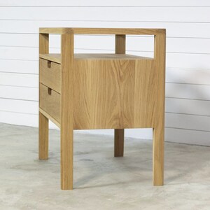 Puede incluir: Una mesita de noche de madera clara con dos cajones y un estante abierto. La mesita de noche tiene un dise&ntilde;o simple y moderno.