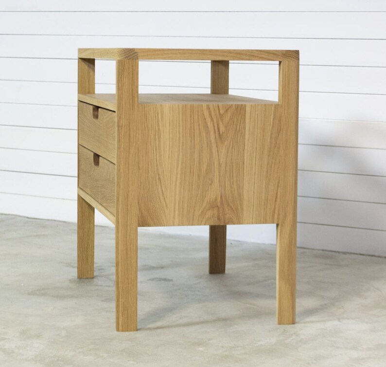 Puede incluir: Una mesita de noche de madera clara con dos cajones y un estante abierto. La mesita de noche tiene un dise&ntilde;o simple y moderno.