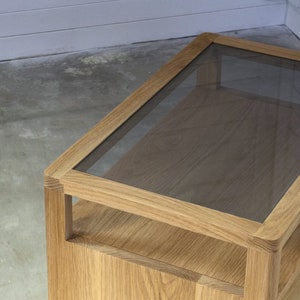 Puede incluir: Una mesa auxiliar de madera con una tapa de vidrio. La mesa tiene un estante debajo de la tapa de vidrio.