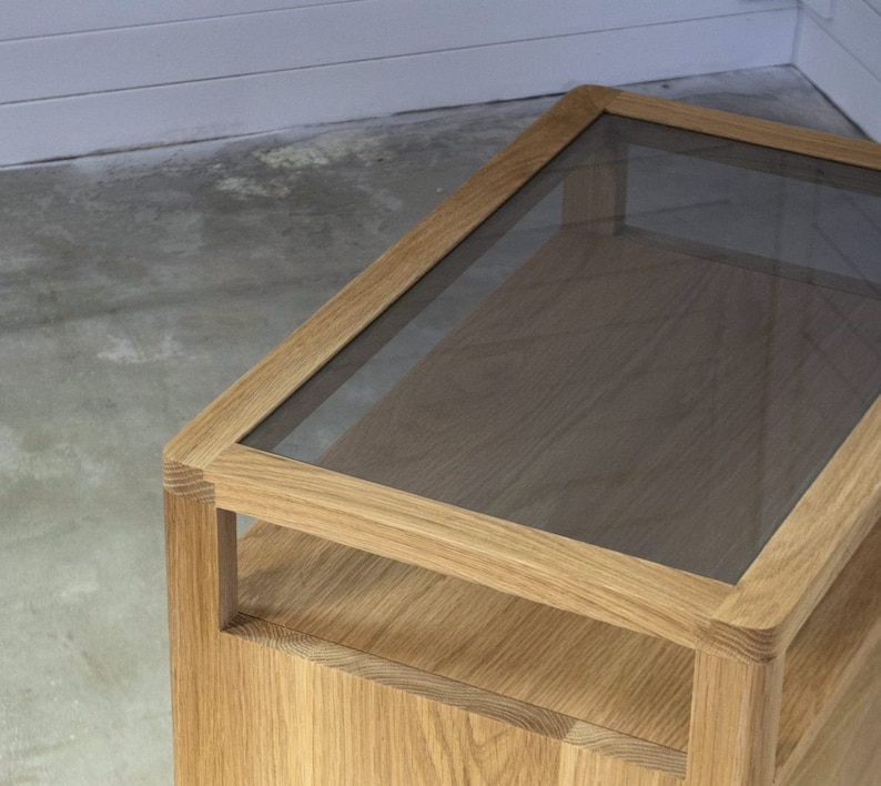 Puede incluir: Una mesa auxiliar de madera con una tapa de vidrio. La mesa tiene un estante debajo de la tapa de vidrio.