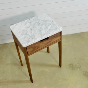 May include: A white marble top nightstand with a single drawer and four wooden legs. The nightstand is made of walnut wood.