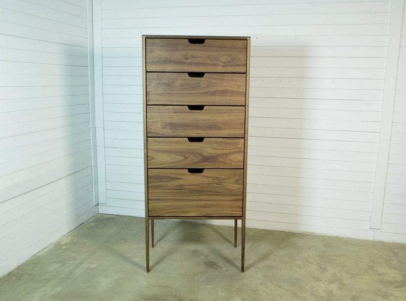 May include: A tall wooden dresser with six drawers and thin metal legs. The dresser is made of a light brown wood with a natural grain pattern.